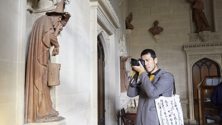 A visitor with a camera up to his face, one eye closed, as he takes a photo of a nearby statue of a figure with a pointy hat and cloak on
